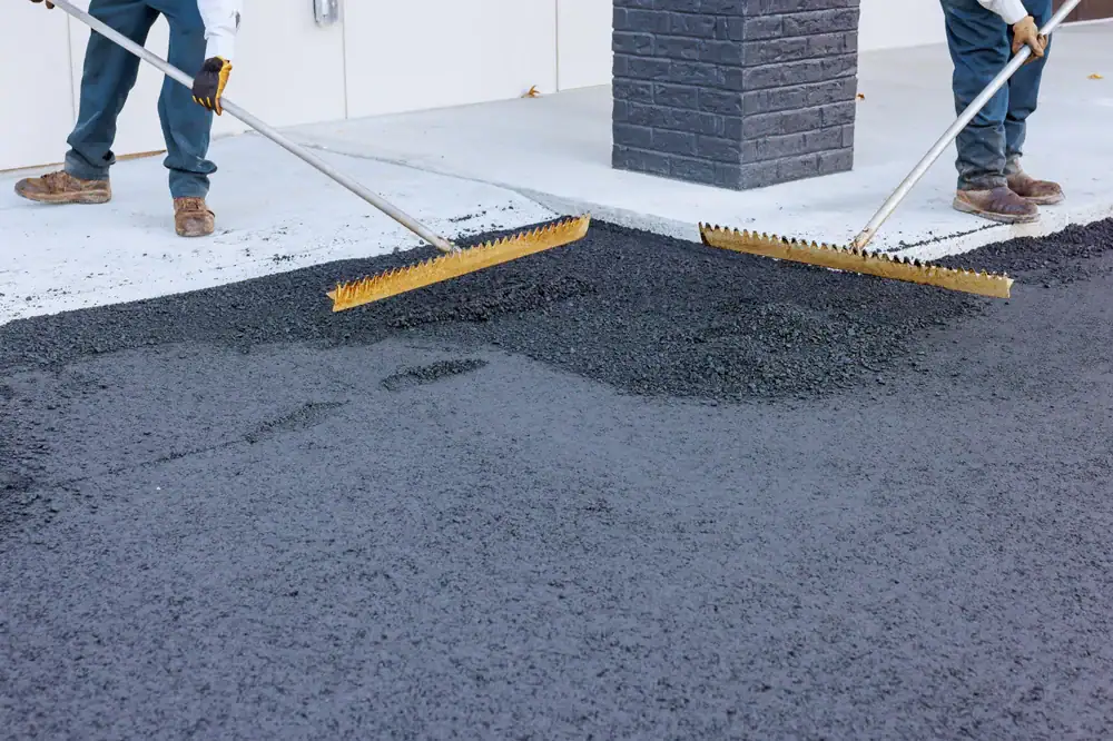 Two workers use large rakes to spread fresh asphalt on a driveway next to a brick column. Only their legs and tools are visible as they work on the paving.