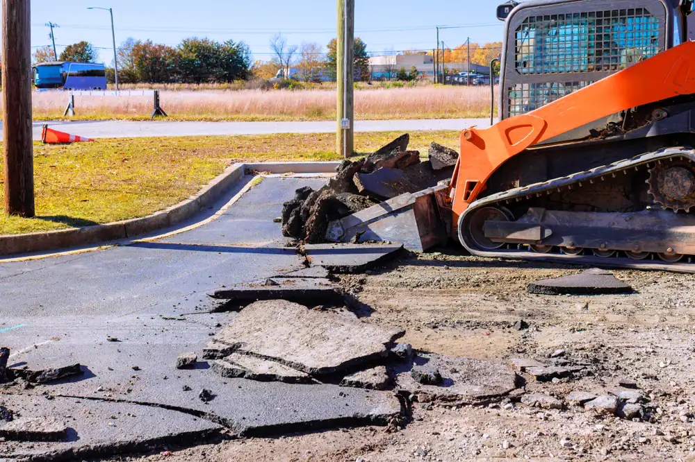 A construction vehicle is breaking up and removing pieces of asphalt from a parking lot, with large chunks of pavement scattered on the ground. Trees and a road are visible in the background.