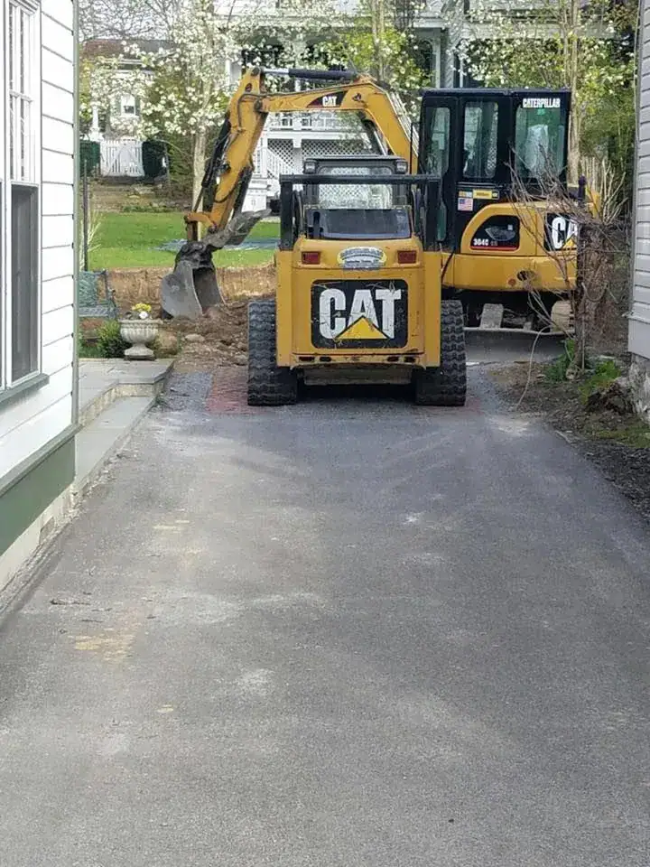 A small yellow CAT construction vehicle is parked on a driveway between two houses, facing another CAT excavator in the background near a grassy yard. Trees with white blossoms are visible behind the machinery.