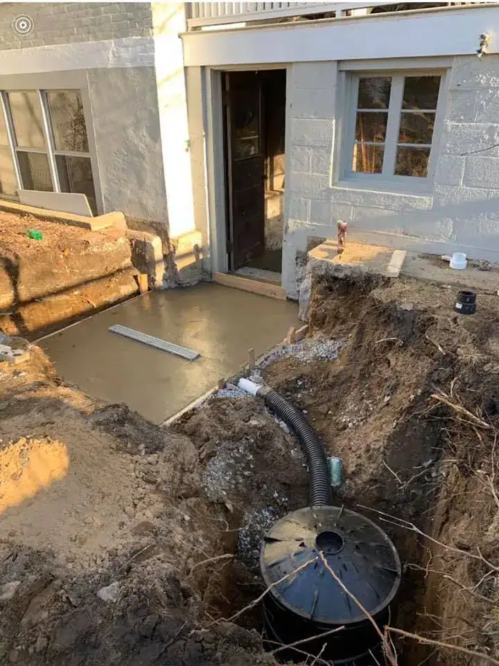 A construction site outside a building shows freshly poured concrete by a door, dug-up earth, a drainage pipe, and a black plastic cylindrical tank partially buried in the ground.