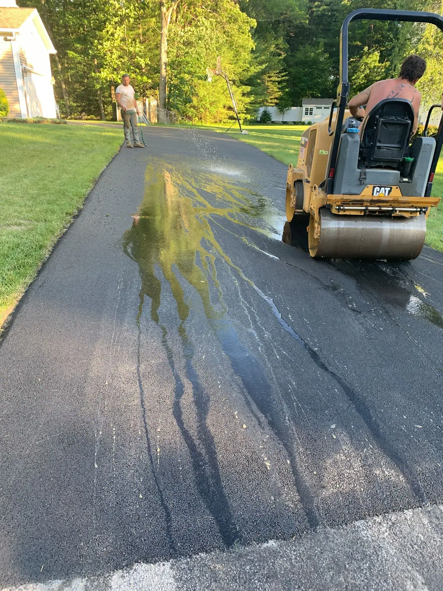 A man operates a steamroller on a freshly paved driveway while another man stands nearby. Wet asphalt or sealant is spread unevenly across the dark surface, creating shiny streaks. Green grass lines both sides of the driveway.