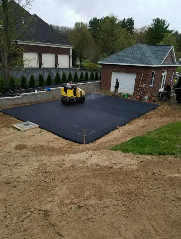 A person operates a small yellow steamroller on freshly paved black asphalt in front of a brick garage, while two other people stand nearby. The area is surrounded by grass, dirt, and a house in the background.