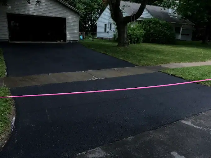 A freshly paved black asphalt driveway leading to a garage, with a pink caution tape across the entrance. Green grass and houses are visible in the background.
