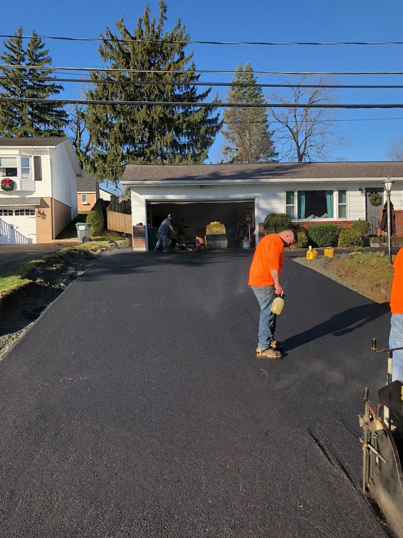 Workers in orange shirts are resurfacing a driveway in front of a suburban house with a garage, using asphalt and construction tools. One worker smooths the surface while others work near the garage. It is a sunny day.