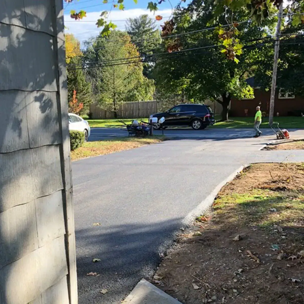 Two workers in safety vests stand near a freshly paved driveway that abruptly ends before reaching the street, leaving a gap. A black SUV and a white car are parked nearby under trees on a sunny day.