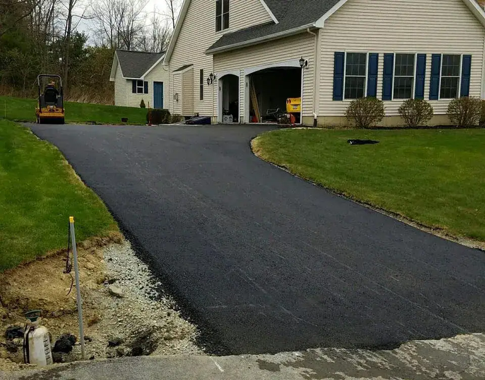 Freshly paved asphalt driveway leading to a beige house with blue shutters, an attached garage, and neatly trimmed green lawns on both sides. Some construction equipment and gravel are visible near the driveway’s edge.