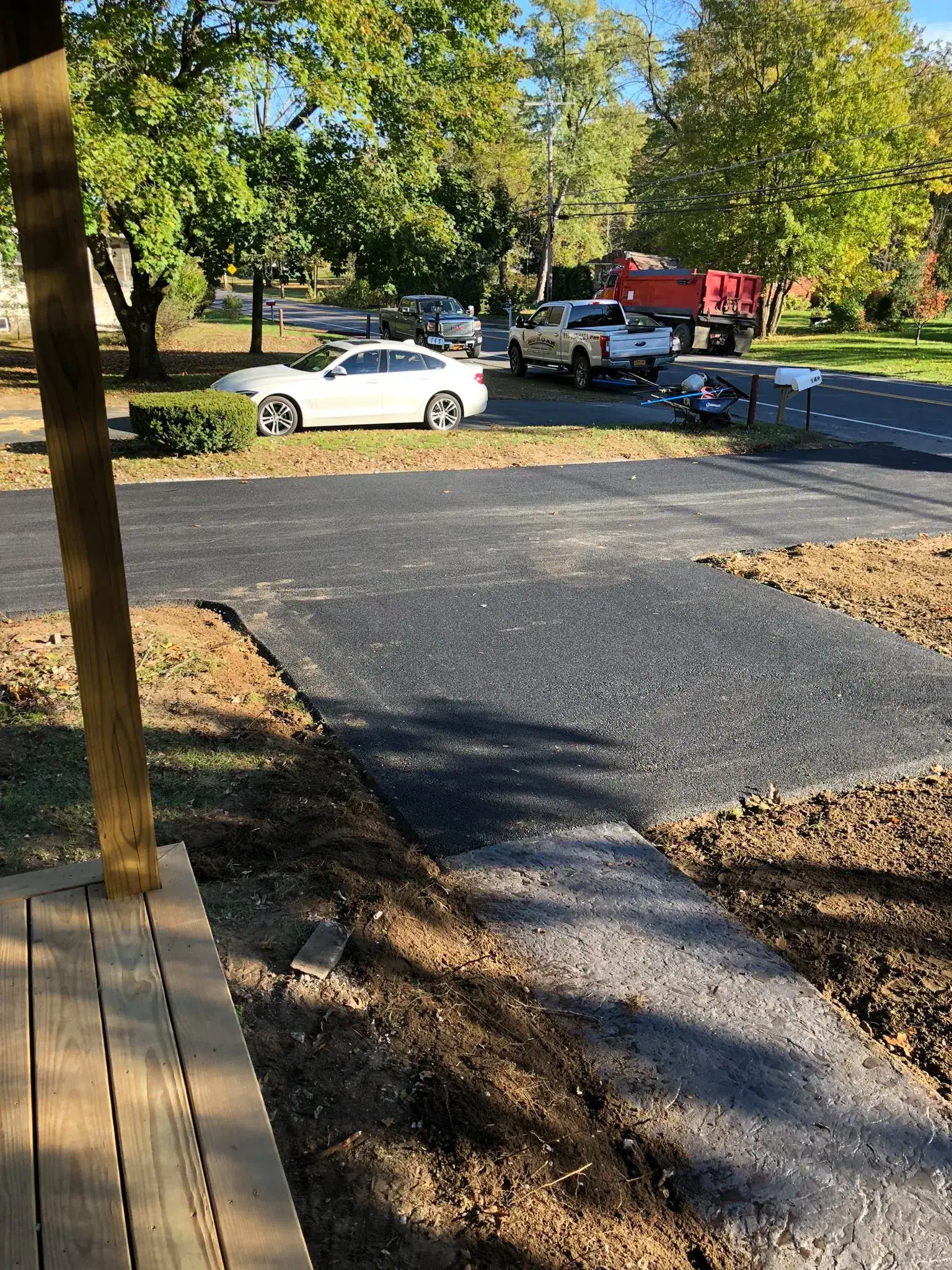 View from a porch showing a newly paved driveway connecting to a street. Several vehicles, including a white car, a white pickup truck, and a red truck, are parked nearby. Trees and mailboxes line the street.