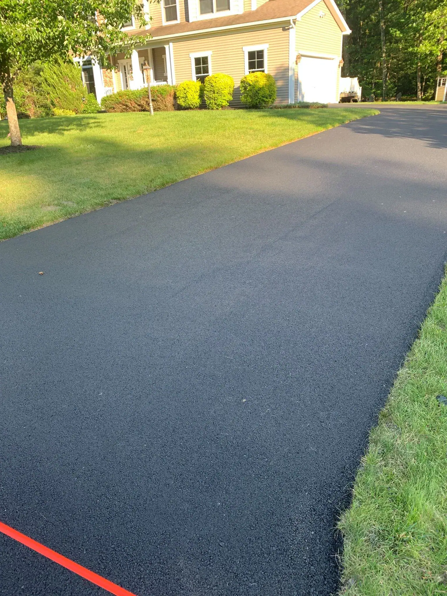 A newly paved black asphalt driveway leads to a beige house with white trim, surrounded by green grass, bushes, and trees, on a sunny day. A red line is visible at the edge of the driveway.