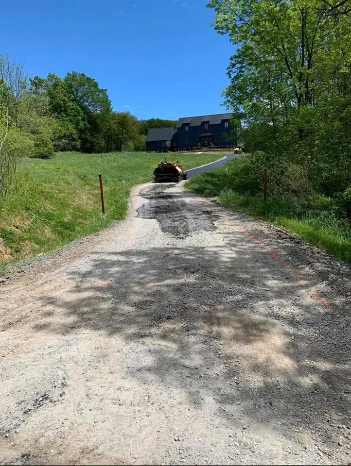 A gravel driveway leads uphill toward a house surrounded by trees and grass under a clear blue sky. A pile of logs sits on the driveway near the house.