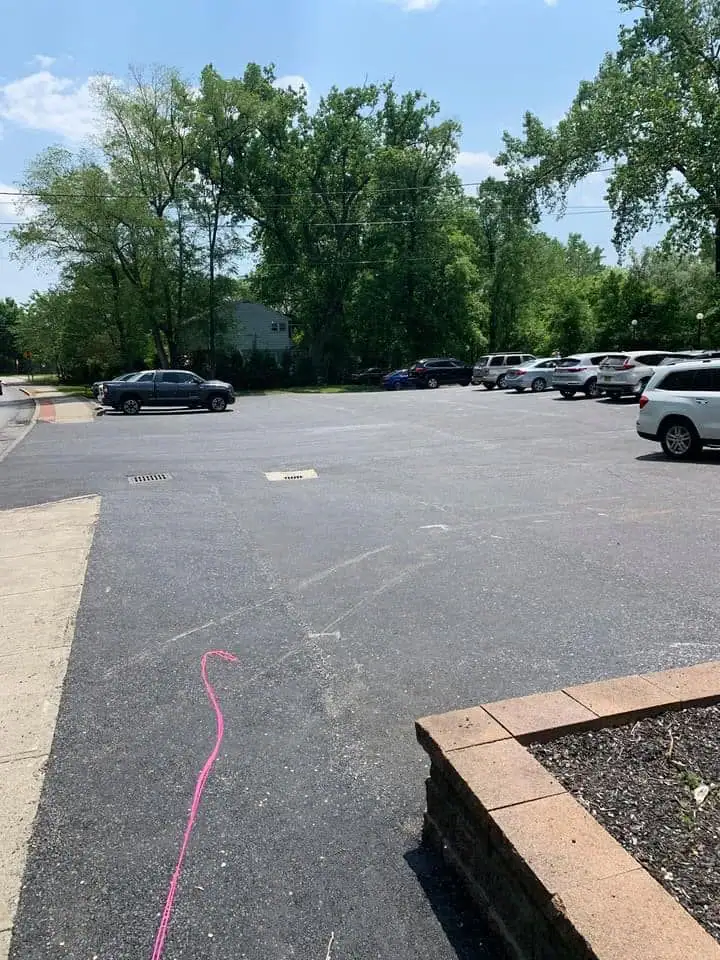 A parking lot with several cars parked on the right, bordered by a sidewalk and a raised flower bed. A pink string lies on the ground, extending from the foreground toward the lot. Green trees and a house are in the background.
