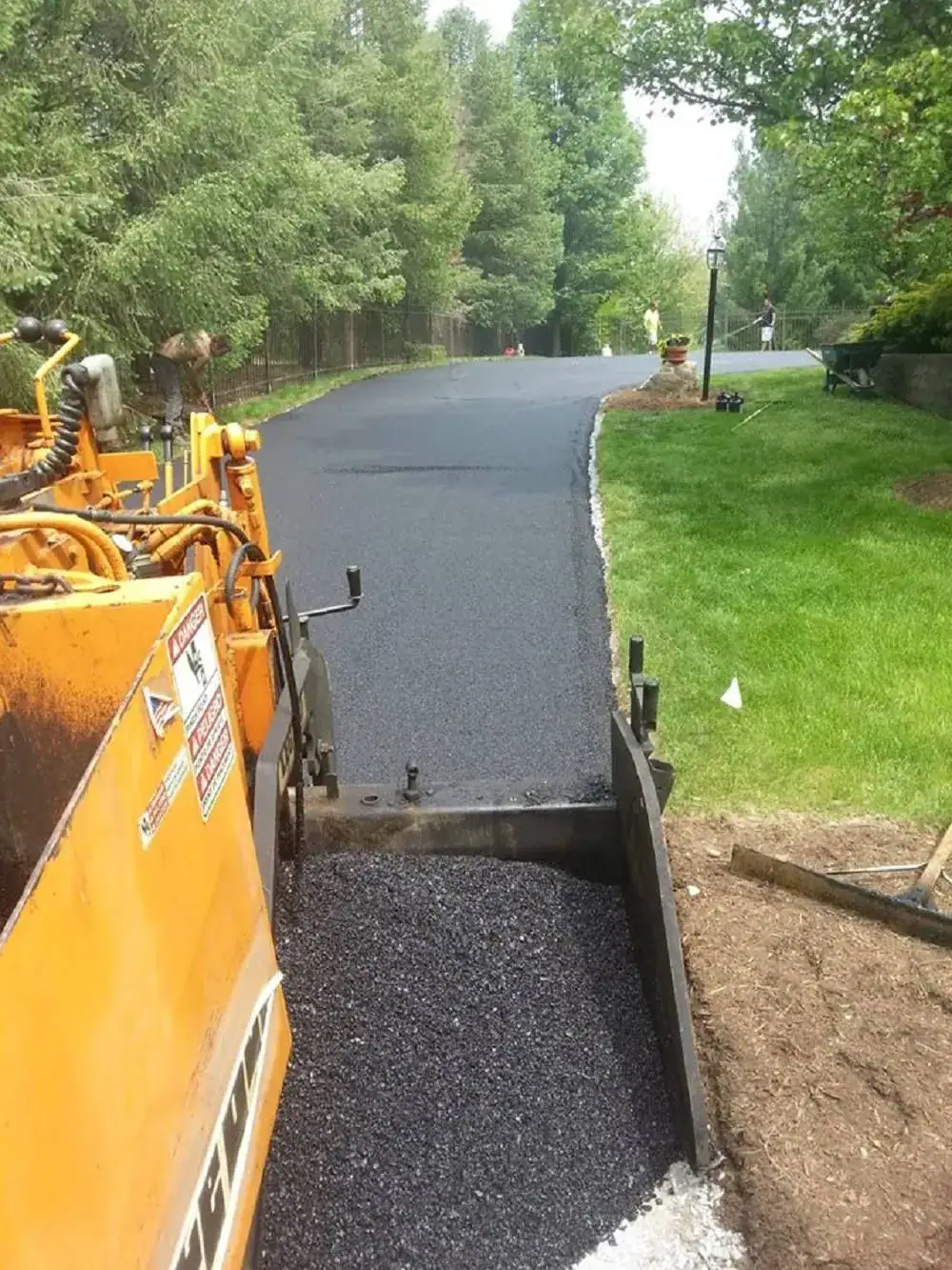 A yellow paving machine lays fresh black asphalt on a driveway surrounded by green grass and trees on a clear day.