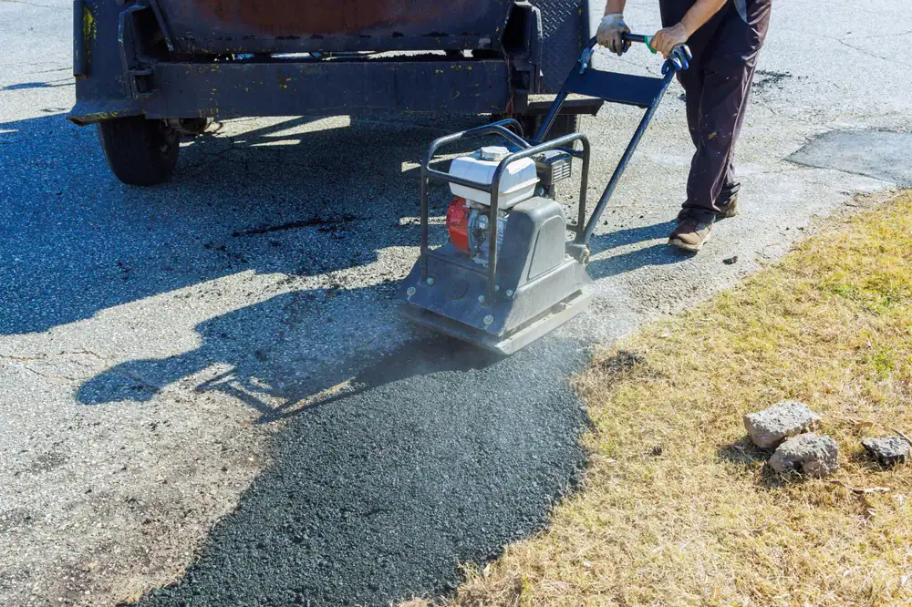 A worker uses a vibrating plate compactor to press down fresh asphalt on a road surface near a patch of grass, with paving equipment visible in the background.
