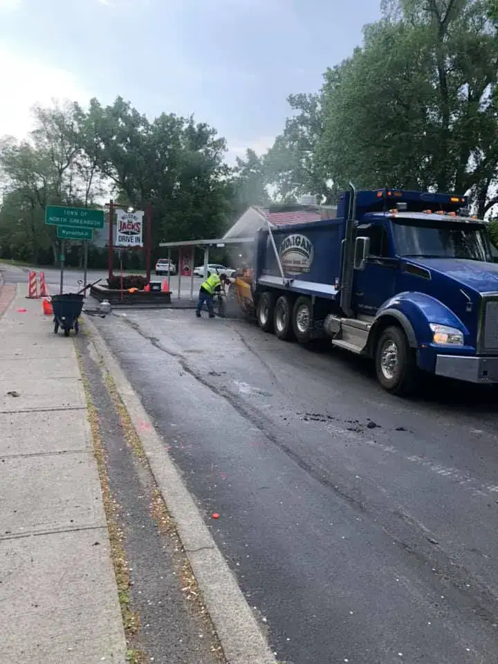 Two workers repair a street beside a large blue dump truck. Steam rises as they work. Nearby, there's a green street sign and a Jack's Drive-In sign. Trees and cones line the background.