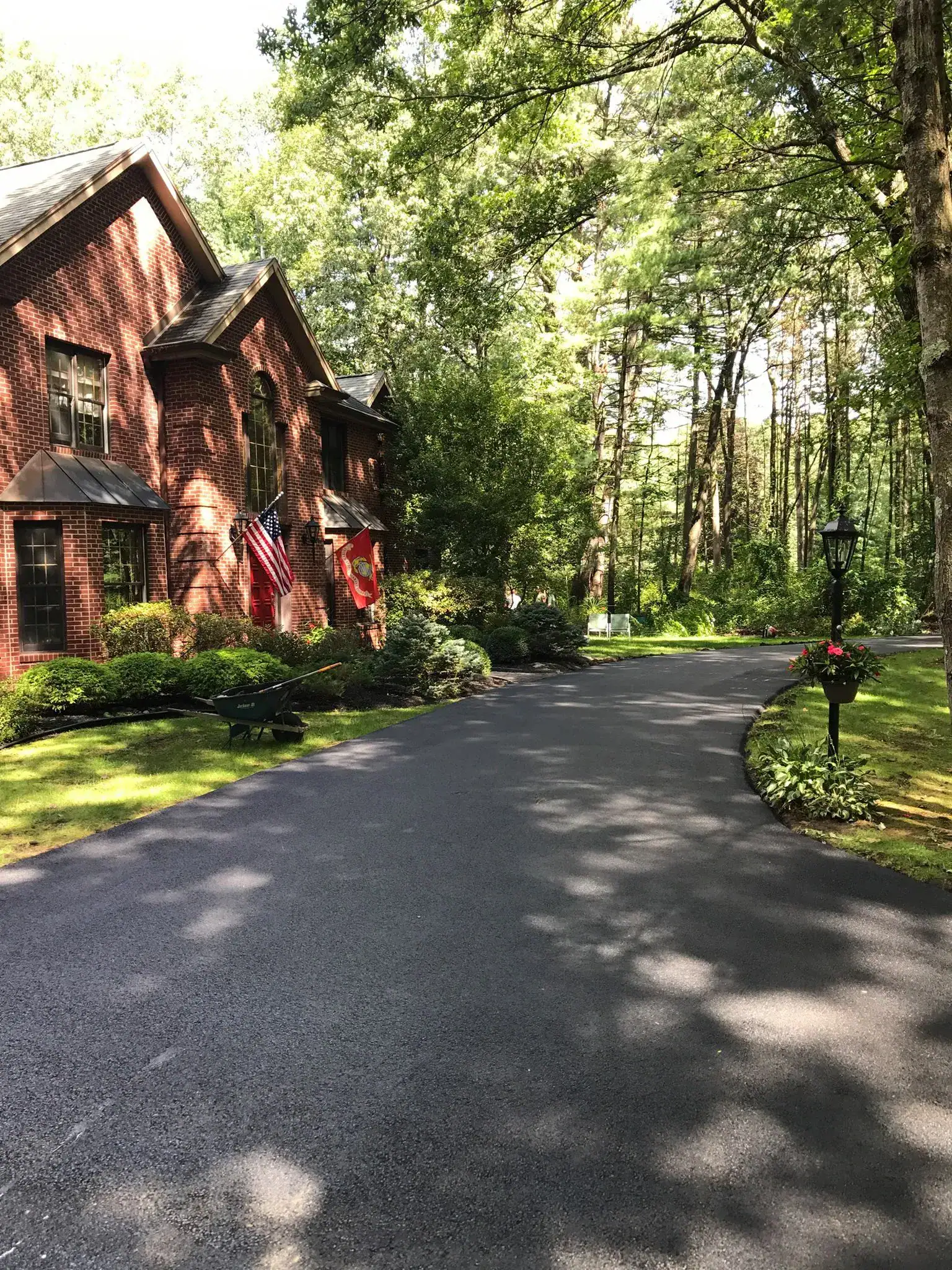 A smooth, newly-paved driveway curves past a brick house with American and Marine Corps flags, surrounded by lush trees, green lawn, bushes, and a black lamppost with hanging flowers. Sunlight filters through the branches.